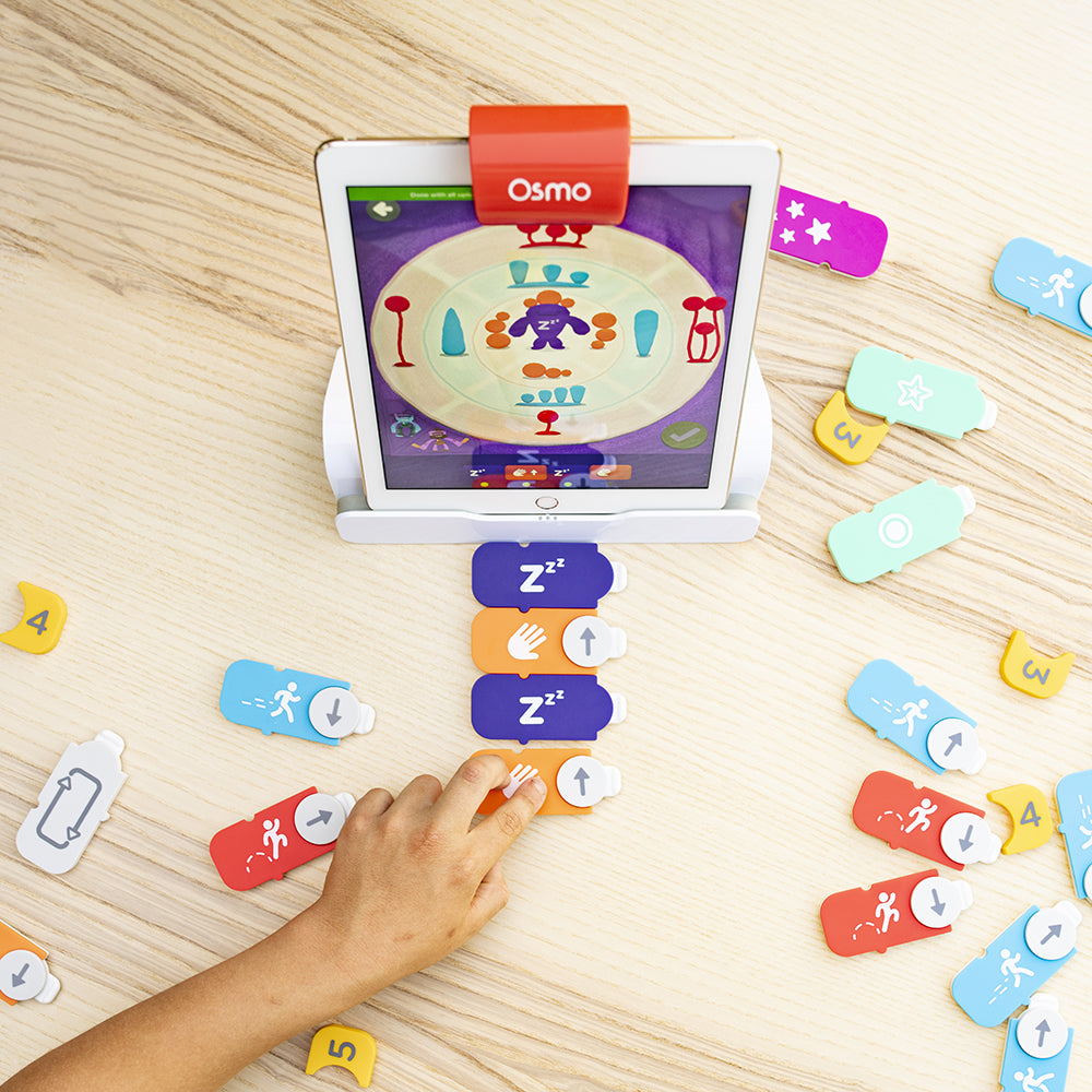 A hand arranging a sequence of coding blocks while Awbie's adventure plays on the iPad